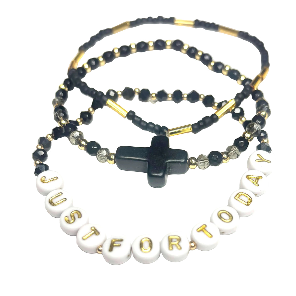 A beaded bracelet stack with white letter beads spelling 'JUST FOR TODAY' in gold, a black cross pendant, and black faceted beads with gold accents on a white background.